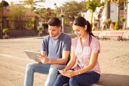 Young couple sitting in a town square on a bench looking at a tablet and a cell phone while dressed casually in jeans and t-shirtsの写真素材