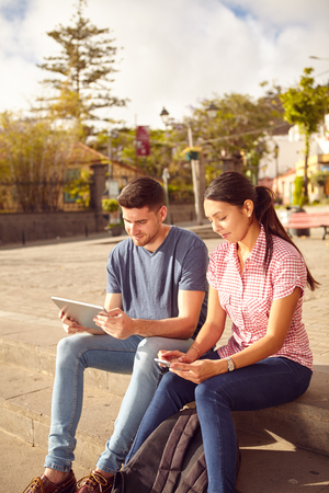 Young couple sitting in a town square on a bench looking at a tablet and a cell phone while dressed casually in jeans and t-shirtsの写真素材