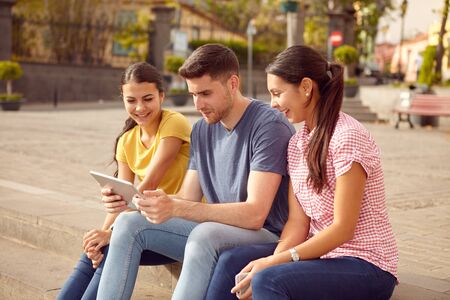 Three young people sitting in a town square on a step looking at a tablet smiling while dressed casually in jeans and t-shirtsの写真素材