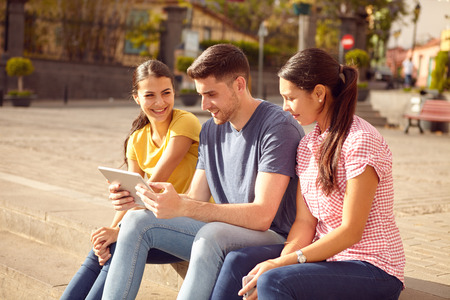 Three young people sitting in a town square on a step looking at a tablet smiling while dressed casually in jeans and t-shirtsの写真素材