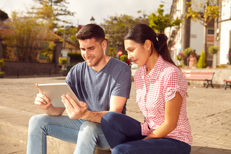 Young couple sitting in a town square on a bench in the sunshine looking at a tablet while dressed casually in jeans and t-shirtsの写真素材