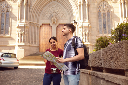 Young tourist couple on vacation with a map, a backpack, and a camera while dressed casually in jeans and t-shirts with a cathedral behind themの写真素材