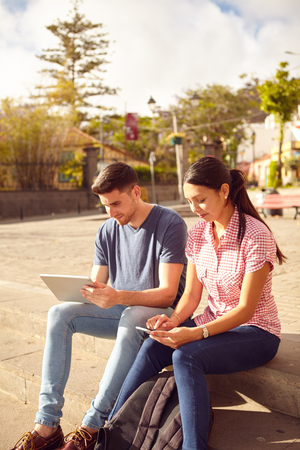 Young couple sitting in a town square on a bench looking at a tablet and a cell phone while dressed casually in jeans and t-shirtsの写真素材