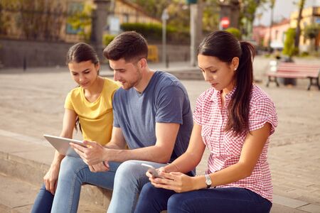 Three young people sitting in a town square on a step looking at a tablet and a cell phone while dressed casuallyの写真素材
