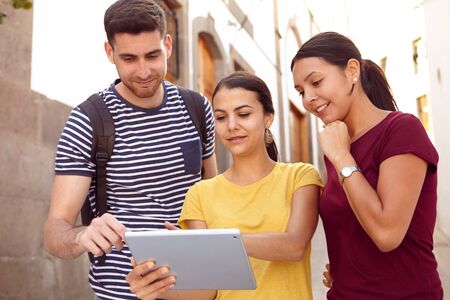 Young tourist couple on vacation with a backpack looking at a tablet with a friend in a narrow street while dressed in casual clothesの写真素材