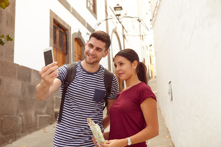 Young tourist couple on vacation with a backpack, a map taking a selfie in a narrow street while dressed casually in jeans and t-shirtsの写真素材