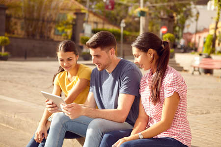Three young people sitting in a town square on a step looking at a tablet smiling while dressed casually in jeans and t-shirtsの写真素材