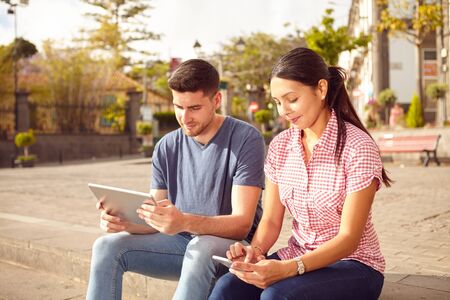 Young couple sitting in a town square on a bench looking at a tablet and a cell phone while dressed casually in jeans and t-shirtsの写真素材