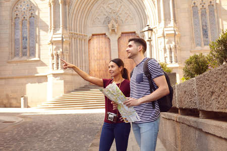 Young tourist couple on vacation with a map, a backpack, and a camera while dressed casually in jeans and t-shirts with a cathedral behind themの写真素材