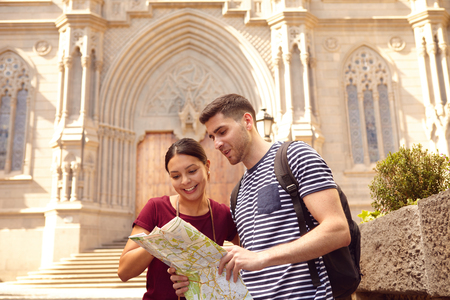 Young tourist couple on vacation with a backpack, looking at a map while dressed casually in jeans and t-shirts with a cathedral behind themの写真素材