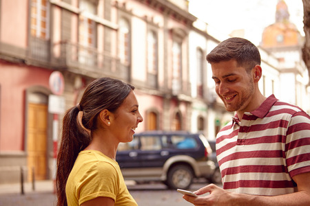 Young couple looking at the cell phone smiling happily while facing each other dressed casually t-shirts with buildings behind themの写真素材