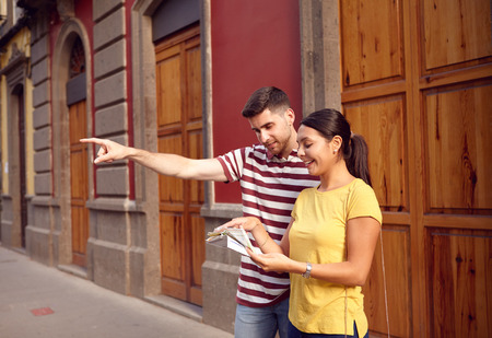 Young couple studying a map while pointing to their front with happy smiles and dressed casually in t-shirts with old buildings behind themの写真素材