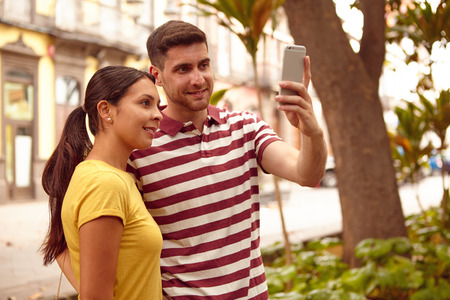 Young couple looking at the cell phone smiling happily while taking a selfie dressed casually in t-shirts in a tree lined streetの写真素材
