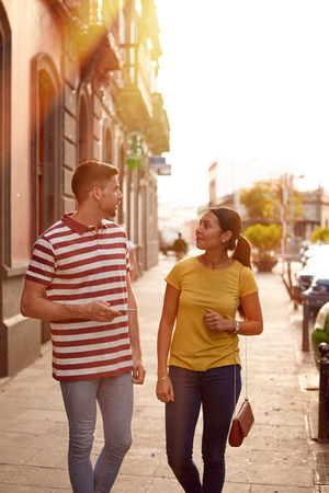 Young couple looking at each other talking happily while facing each other dressed casually t-shirts with buildings behind themの写真素材