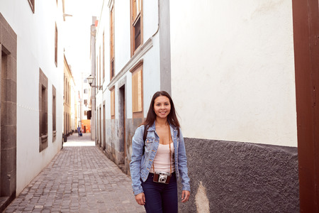 Pretty young girl posing in a narrow paved street looking at the camera with old gray and white buildings behind her in casual clothingの写真素材