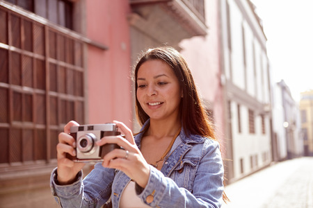 Pretty young girl looking at a picture in a narrow paved street with old pink buildings behind her in casual jeans and denim jacketの写真素材