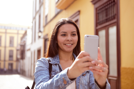 Pretty young girl taking pictures smiling on her cell phone in a narrow street with old yellow buildings behind her in casual clothingの写真素材