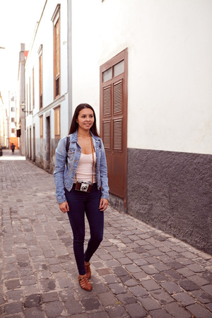 Pretty young girl strolling down a narrow paved street with old gray and white buildings behind her in casual clothingの写真素材