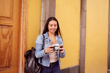 Pretty young girl leaning against a gray pillar looking at her camera with old yellow buildings behind her in blue jeans and a denim jacketの写真素材