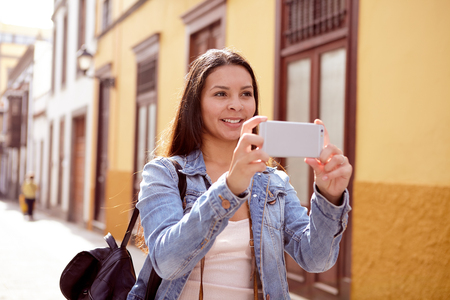 Pretty young girl looking up to take a picture with her cell phone in a narrow paved street wearing a denim jacketの写真素材