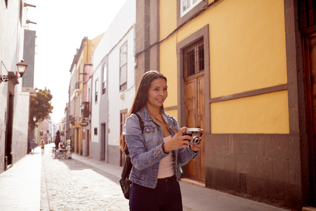 Pretty young girl taking pictures in a narrow paved street with old buildings wearing casual blue jeans and a denim jacketの写真素材
