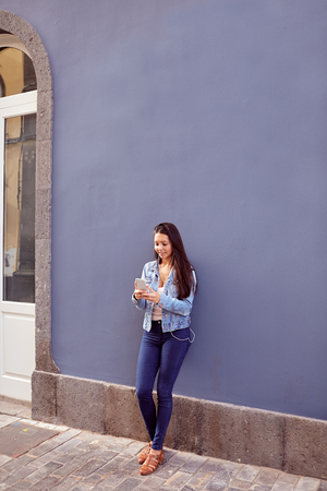 Cute young girl looking at her cell phone while leaning against a blue wall smiling and her hair loose in jeans and denim jacketの写真素材