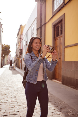 Pretty young girl taking a picture in a narrow paved street with old buildings behind her in blue jeans and denim jacketの写真素材