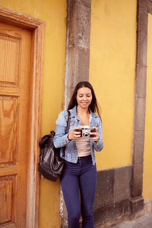 Pretty young girl leaning against a gray pillar smiling at her camera with old yellow buildings behind her in casual jeans and denim jacketの写真素材