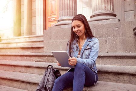 Pretty young girl looking at her tablet while sitting on the steps of an old building smiling wearing her hair loose and casual clothingの写真素材