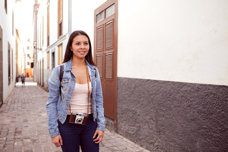 Pretty young girl standing in a narrow paved street with old gray and white buildings behind her in casual clothingの写真素材
