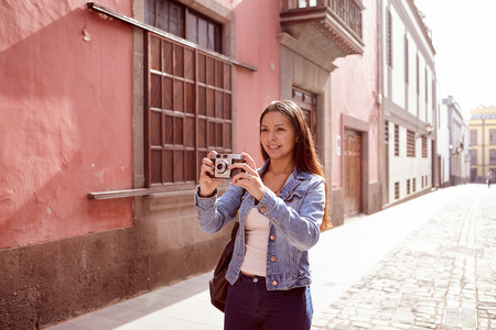 Smiling young girl taking a picture in a narrow paved street with old pink buildings behind her in casual jeans and denim jacketの写真素材