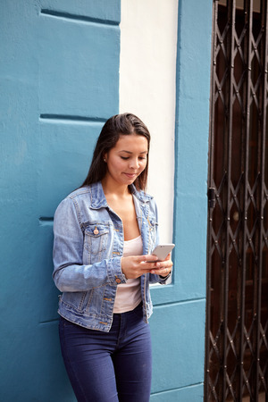 Pretty young girl typing on her cell phone while leaning against a blue wall with a white niche and smiling in casual clothingの写真素材