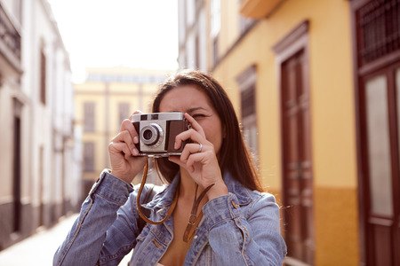 Cute young girl taking a picture in a narrow paved street with old yellow buildings behind her in a casual denim jacketの写真素材