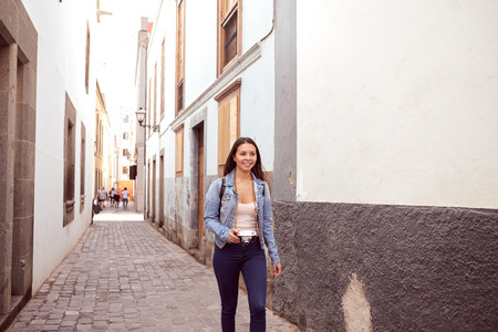Pretty young girl hurrying down a narrow paved street looking at the camera with old gray and white buildings behind her in casual clothingの写真素材