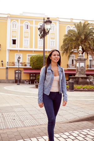 Pretty young girl strolling and looking ahead with a toothy smile and wearing casual clothing and a backpack, old buildings behind herの写真素材