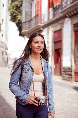Pretty young girl strolling down a narrow street with a camera wearing her hair loose and casual clothing with a narrow street in her backgroundの写真素材