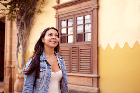 Pretty young girl standing, looking up while wind is blowing her long hair back and wearing casual clothing with an old building behind herの写真素材