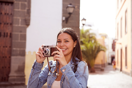 Pretty young girl taking pictures and looking at the camera wearing her long hair loose and casual clothing with a narrow street in her backgroundの写真素材