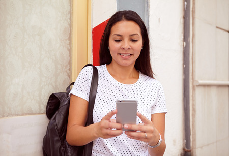 Pretty young girl smiling at her cell phone while standing in front of a pillar, wearing her hair loose and a spotted t-shirtの写真素材