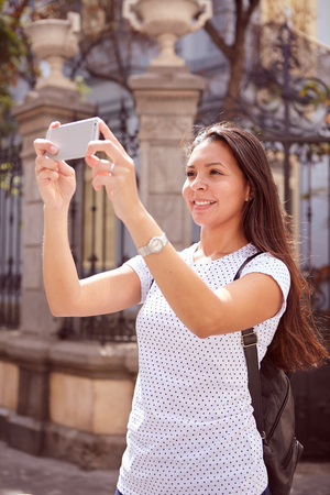 Cute young girl taking pictures with her cell phone while standing in front of old gates, wearing her hair loose and a spotted t-shirtの写真素材