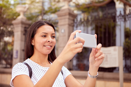 Pretty young girl smiling at her cell phone while standing in front of old buildings, wearing her hair loose and a spotted t-shirtの写真素材