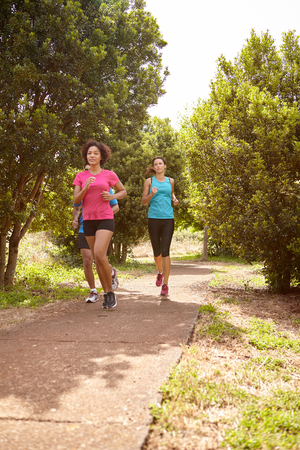 Three young runners on a paved jogging trail in daylight surrounded by trees and bushes in the late morning wearing t-shirts and black pantsの写真素材