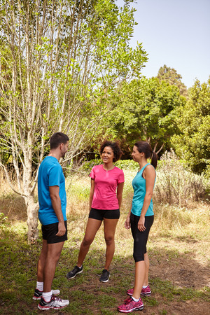 Three young joggers taking a break in daylight surrounded by trees and bushes in the late morning wearing t-shirts and black pantsの写真素材