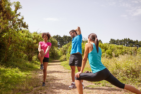A male and two female friends doing some stretching on a jogging trail surrounded by trees in the late morning wearing t-shirts and black pantsの写真素材