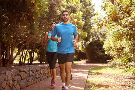 A guy and two girls running on a paved running trail with trees and a stone wall behind them wearing t-shirts and black pantsの写真素材