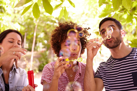 Three funloving friends blowing bubbles and smiling in the dappled afternoon sunshine with some trees around them wearing casual clothingの写真素材