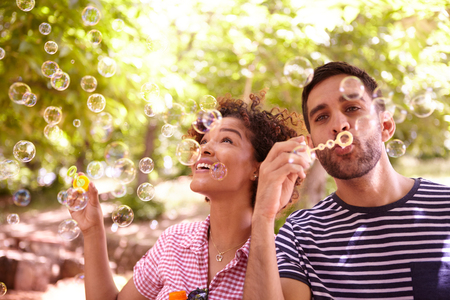 Two young friends joyfully blowing bubbles and smiling in the dappled afternoon sunshine with some trees around them wearing casual clothingの写真素材