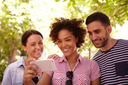 Three young people looking and smiling at the cell phone in the shade of some trees wearing casual...