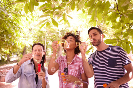 Three young people blowing some bubbles in the dappled afternoon sunshine with some trees around them wearing casual clothingの写真素材