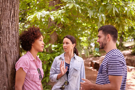 Two girl friends and a guy chatting in the dappled afternoon sunshine while one girl leans against a tree wearing casual clothingの写真素材